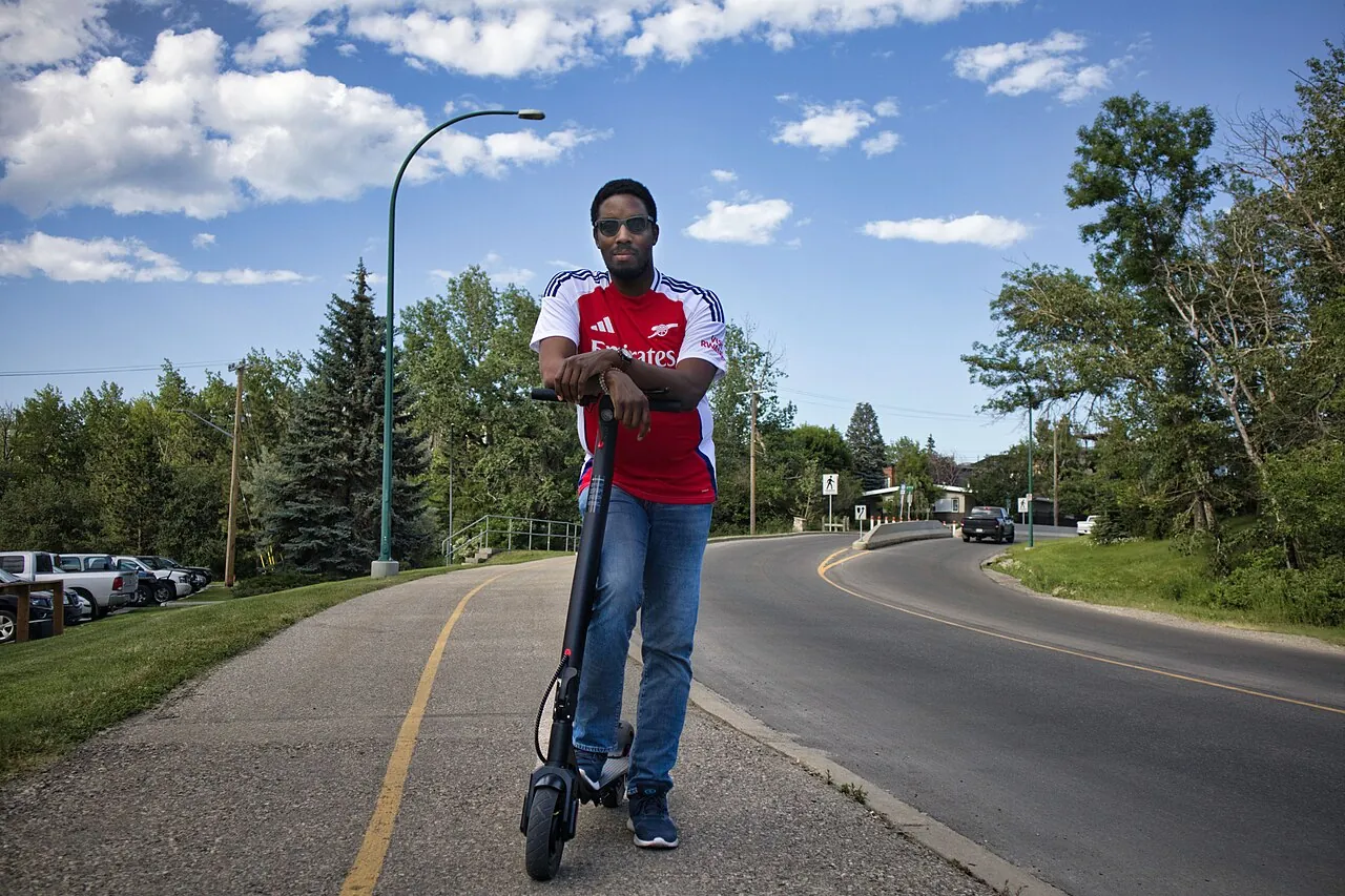 A person standing with a commuter scooter beside a paved path.
